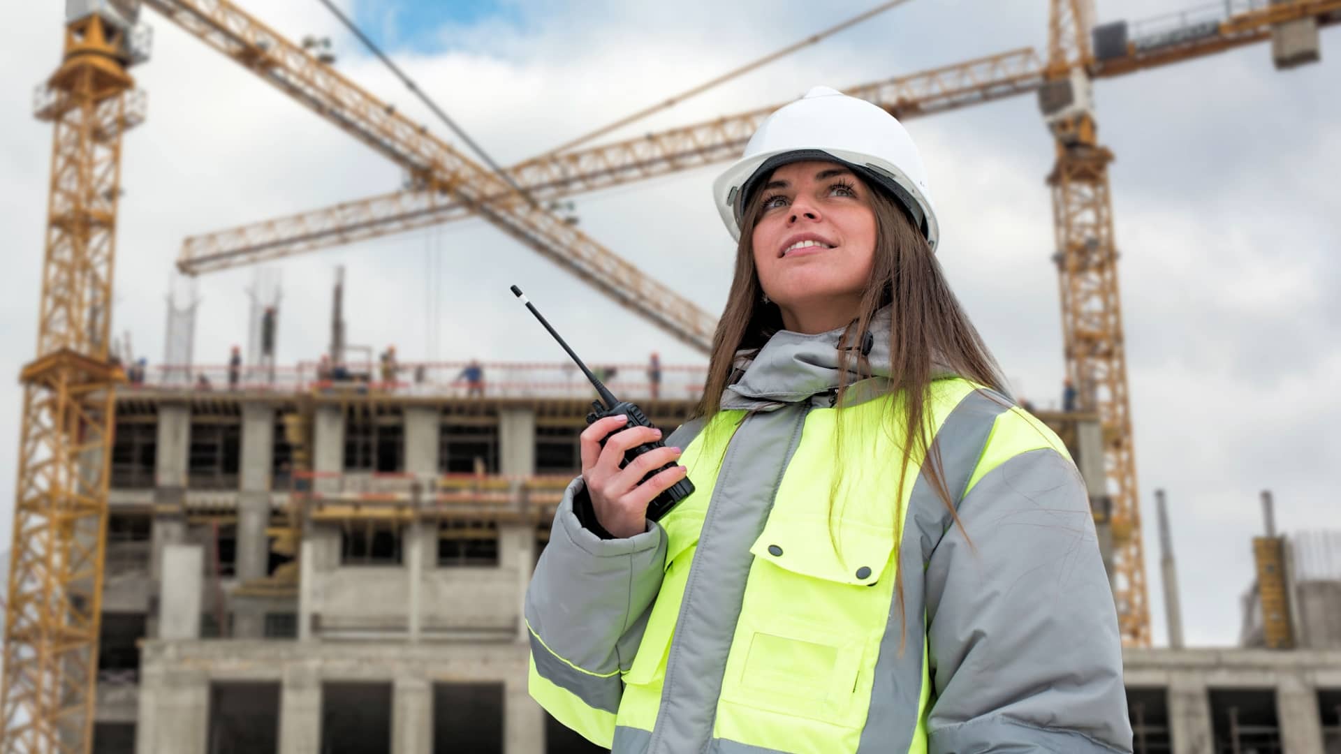 Construction supervisor in a white hard hat and hi-vis jacket holding a two-way radio on a building site with tower cranes