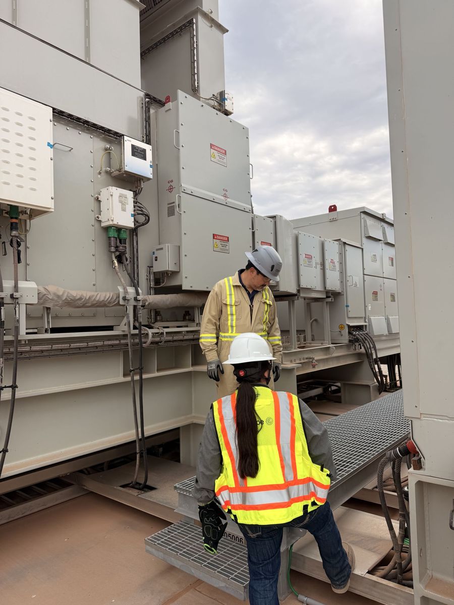 Two workers in hard hats and hi-vis vests on a metal walkway between large equipment cabinets at a power plant