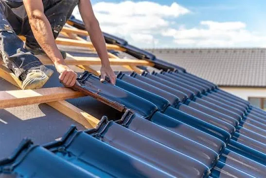 Roofer's hands setting dark blue clay tiles onto wooden battens on a sunny roof